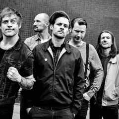 Fünf Männer posieren vor einer Ziegelwand, wie Punkbands der 80er Jahre. Sie tragen Jeansjacken, Bomberjacken und karierte Hemden.