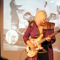 Eine deutsche Gitarristin mit blondem Haar spielt auf einer E-Gitarre vor einer Projektion einer Jazzband.
