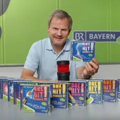 Mann präsentiert CDs mit dem Titel "One Hit Wonder". Viele weitere CDs dieser Reihe sind auf dem Tisch aufgereiht.