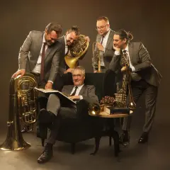 Fünf Musiker mit Blasinstrumenten, darunter Tuba, Horn und Trompete, umringen einen Mann im Sessel, der ein Buch liest.