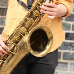 Ein Musiker spielt ein goldenes Saxophon vor einer Ziegelwand. Hier ist ein Saxophon Steckbrief: ein Blasinstrument mit Klappen und einem Mundstück.