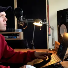 Ein junger Mann mit rotem Pullover und Kappe arbeitet an einem Laptop in einem Musikstudio. Er komponiert Musik, die die **lofi bedeutung** von Entspannung und Kreativität verkörpert.