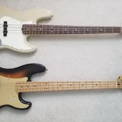 Zwei E-Bässe liegen nebeneinander: oben ein cremefarbener Jazz Bass, unten ein abgenutzter Sunburst Precision Bass.