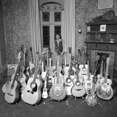 Mann mit vielen Rock n Roll Instrumenten, darunter Akustik- und E-Gitarren, vor einem Fenster.