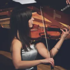 Eine Frau spielt Geige, ihr Bogen erzeugt einen zarten vibrato geige Klang. Im Hintergrund ist ein Klavier zu sehen.