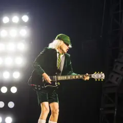 Angus Young von AC/DC spielt auf der Bühne, während die Scheinwerfer leuchten. Seine ikonische Kleidung und die Gitarre lassen an bekannte Lieder denken.