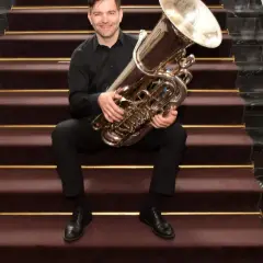 Ein Mann sitzt auf einer Treppe mit einer Tuba. Wer hat die Tuba erfunden? Diese Frage beschäftigt Musiker seit langem.