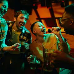 Freunde singen lautstark bei Karaoke mit, halten Getränke und haben sichtlich Spaß. Perfekt für lieder zum mitsingen!