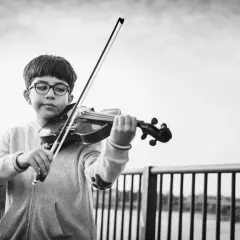 Ein Junge mit Brille spielt konzentriert Geige. Er steht an einer Brüstung, die Musik erklingt im Freien.