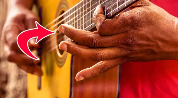 Nahaufnahme der Hände eines Fingerstyle-Gitarristen, der die Saiten einer Akustikgitarre spielt. Ein roter Pfeil zeigt auf die Finger.