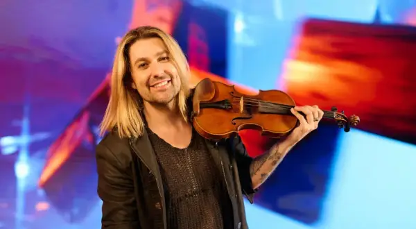 Ein lächelnder Mann mit langen blonden Haaren hält eine Geige. Er könnte der beste Geiger der Welt sein, mit seiner Leidenschaft für Musik.
