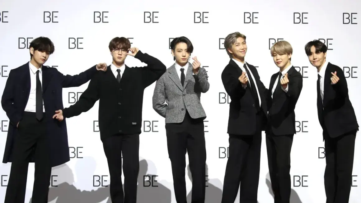 Sechs junge Männer, die wie eine Boyband aussehen, posieren vor einer Wand mit dem Schriftzug "BE".