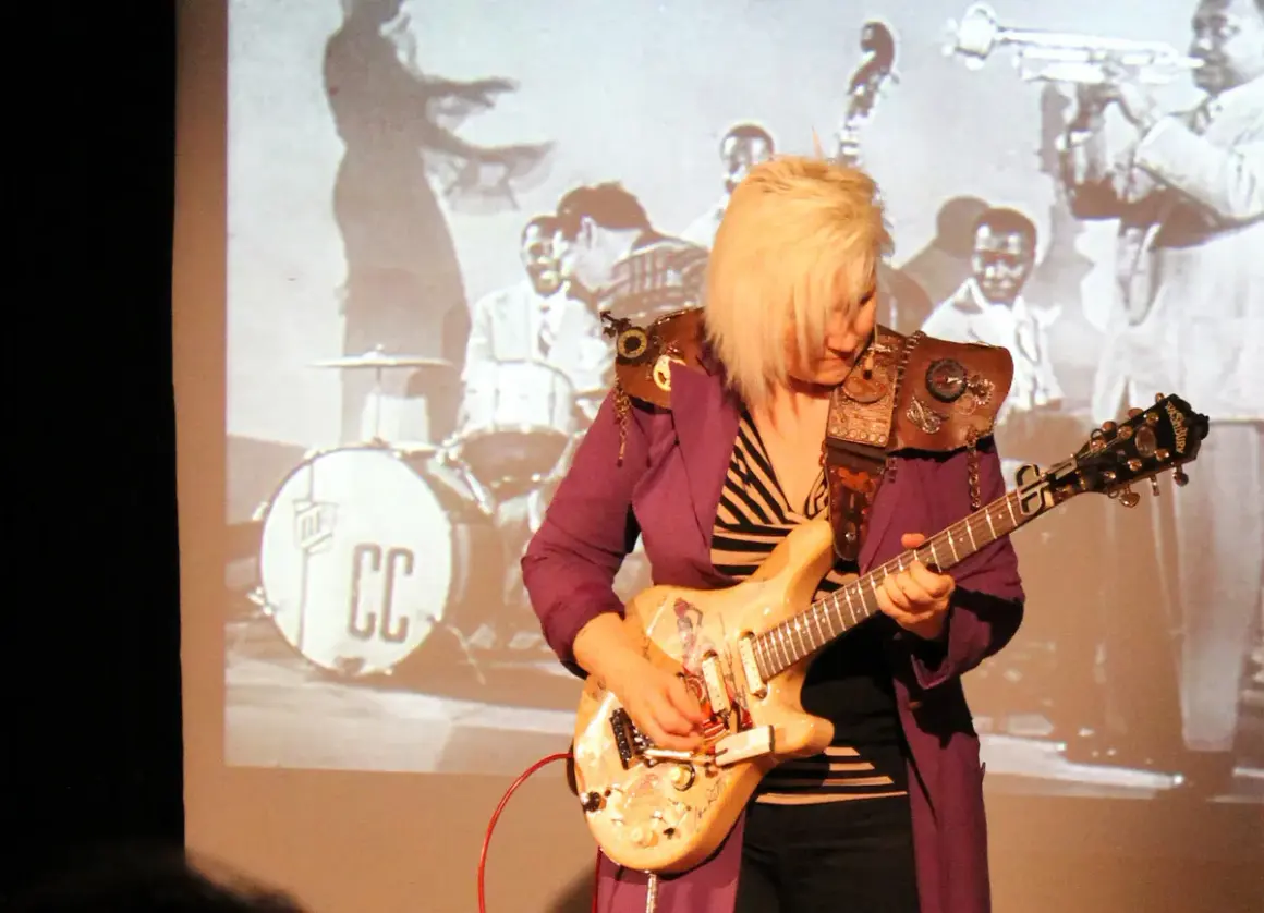 Eine deutsche Gitarristin mit blondem Haar spielt auf einer E-Gitarre vor einer Projektion einer Jazzband.