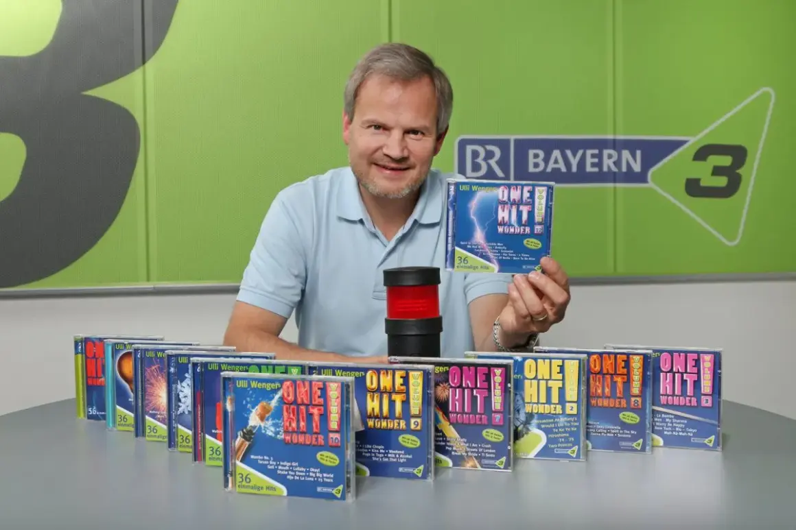 Mann präsentiert CDs mit dem Titel "One Hit Wonder". Viele weitere CDs dieser Reihe sind auf dem Tisch aufgereiht.