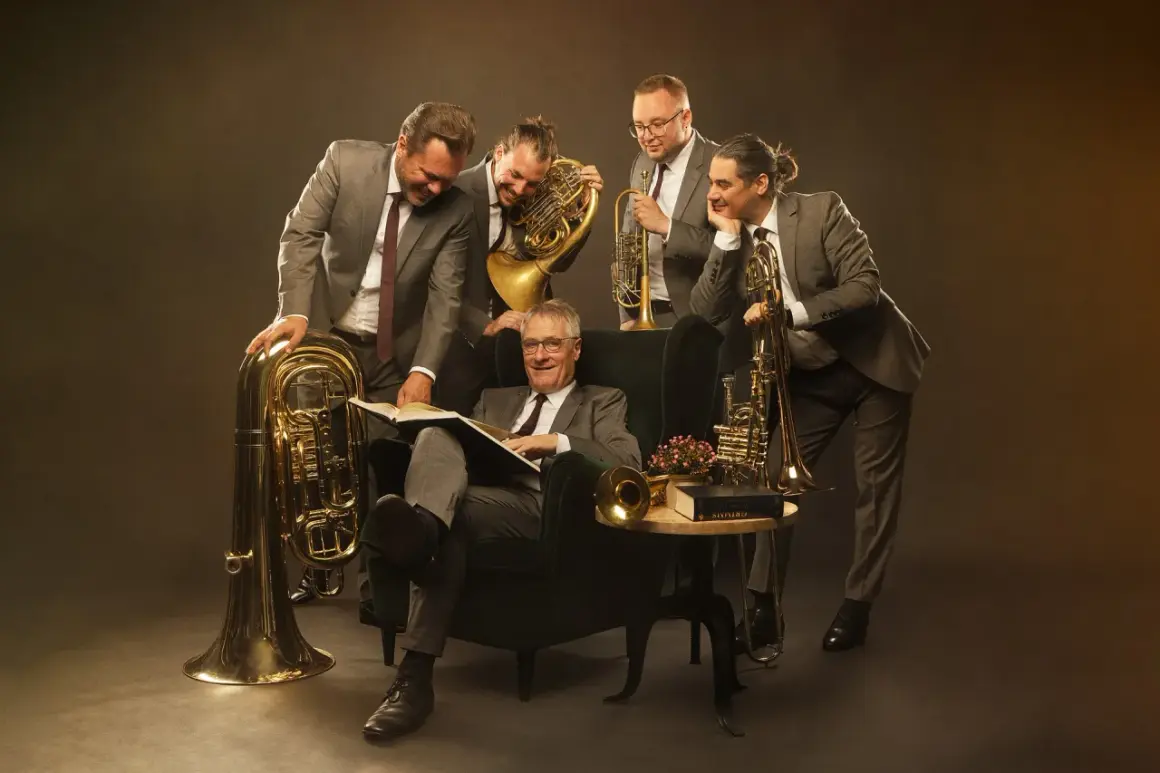 Fünf Musiker mit Blasinstrumenten, darunter Tuba, Horn und Trompete, umringen einen Mann im Sessel, der ein Buch liest.