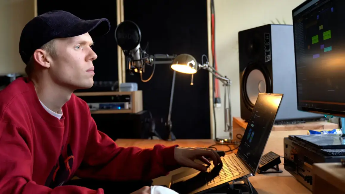 Ein junger Mann mit rotem Pullover und Kappe arbeitet an einem Laptop in einem Musikstudio. Er komponiert Musik, die die **lofi bedeutung** von Entspannung und Kreativität verkörpert.