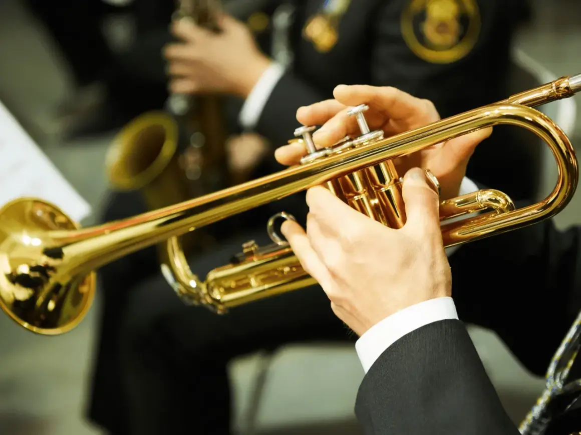 Hände spielen eine glänzende Trompete. Ein Musiker bereitet sich auf sein Referat vor, während im Hintergrund ein Saxophon zu sehen ist.