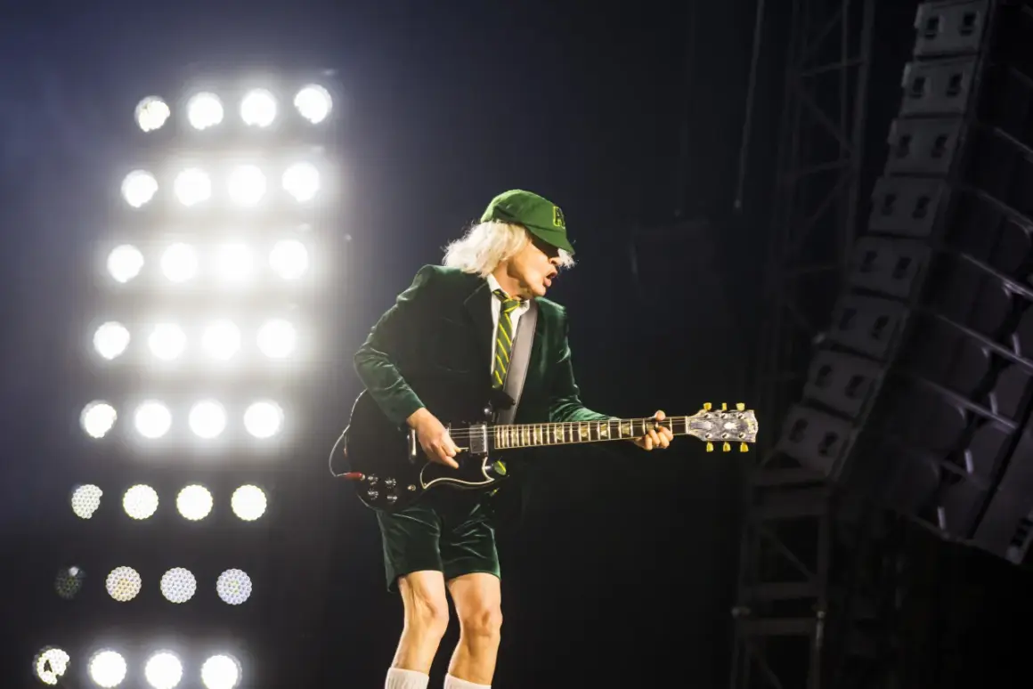 Angus Young von AC/DC spielt auf der Bühne, während die Scheinwerfer leuchten. Seine ikonische Kleidung und die Gitarre lassen an bekannte Lieder denken.