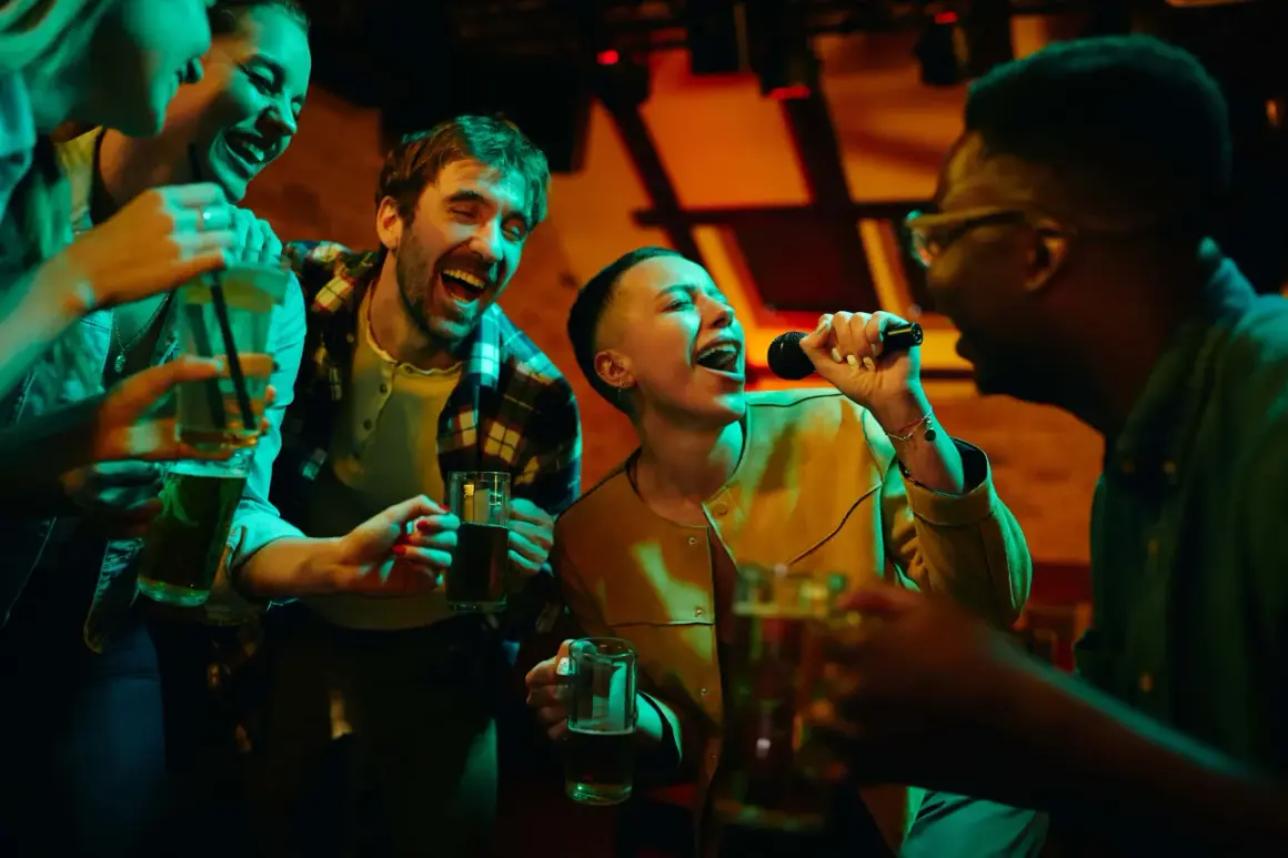 Freunde singen lautstark bei Karaoke mit, halten Getränke und haben sichtlich Spaß. Perfekt für lieder zum mitsingen!