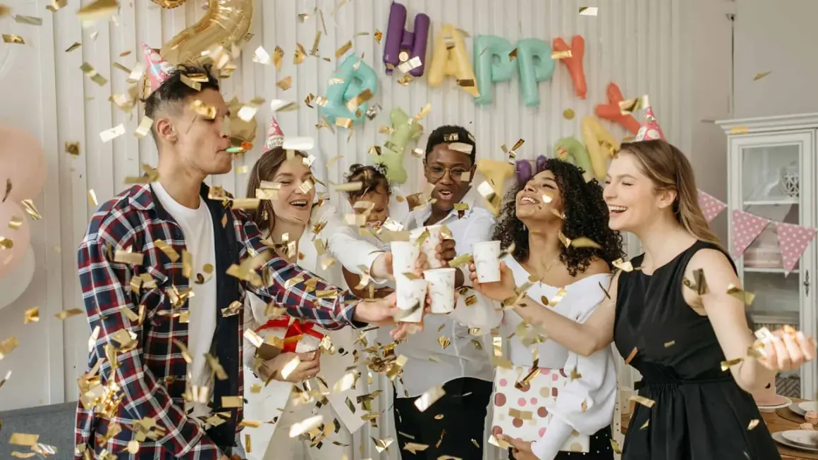 Fröhliche Freunde feiern eine Geburtstagsparty mit Konfetti und Luftballons. Ein "Happy Birthday"-Banner hängt im Hintergrund. Ein tolles Lied für Erwachsene!