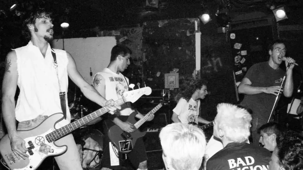 Live-Auftritt einer Punkband in den 80ern. Bassist mit wei&szlig;em Hemd, Gitarrist und S&auml;nger im Vordergrund.
