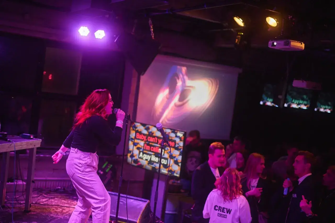 Frau singt mit Mikrofon in einer Karaoke-Bar. Im Hintergrund l&auml;uft ein Songtext auf einem Bildschirm.