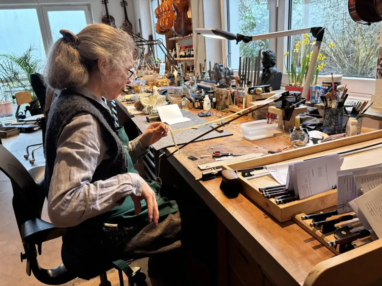 Meisterin bespannt einen Geigenbogen mit Rosshaar. Werkzeuge und Violinen s&auml;umen ihre Werkbank.