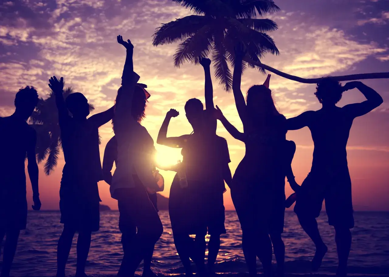 Silhouetten von Menschen tanzen am Strand bei Sonnenuntergang, die Stimmung ist wie bei Ballermann Hits.
