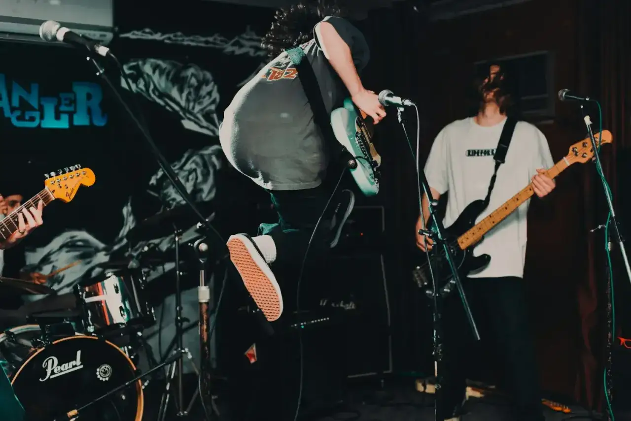 Ein Gitarrist springt auf der B&uuml;hne, ein Bassist spielt im Hintergrund. Energiegeladener Auftritt im Stil des punk 70er.