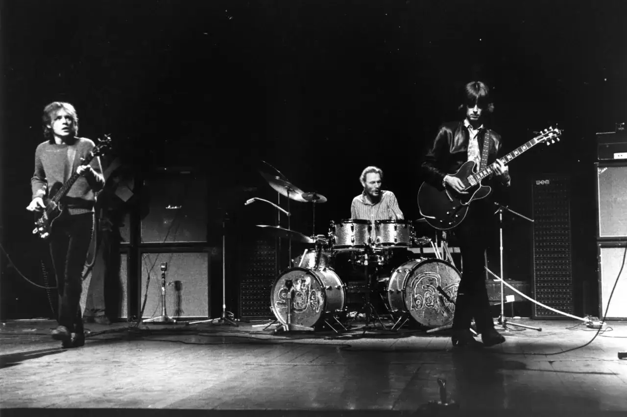 Drei Musiker auf einer Bühne, zwei Gitarristen und ein Schlagzeuger, spielen für eine Menge. Sie sind Teil einer alten Rockband.