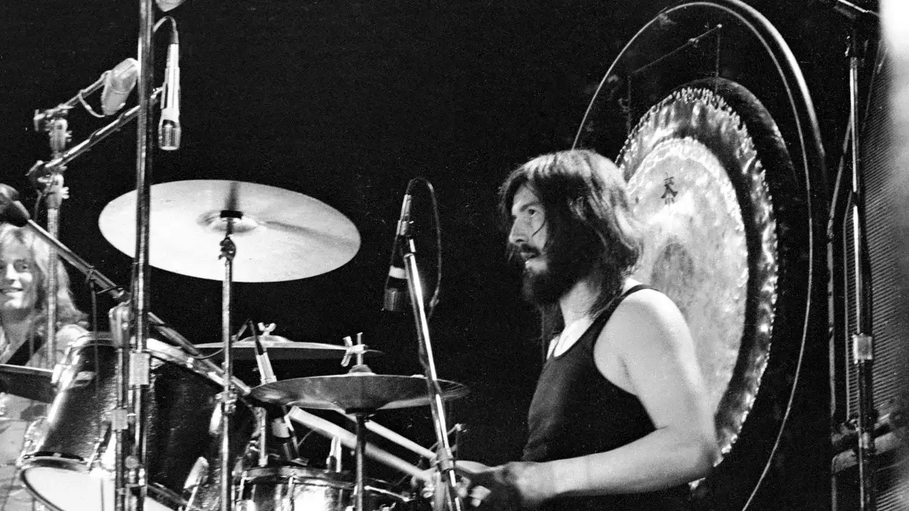 Ein Schlagzeuger mit Bart und schwarzem Tanktop spielt auf einem Schlagzeug, hinter ihm ein Gong. Ein Moment, der an die besten Drum-Solos erinnert.
