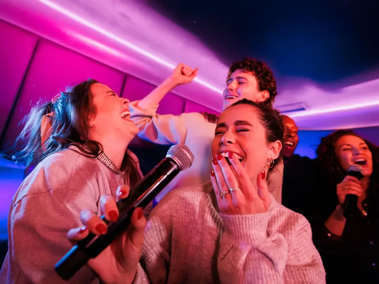 Freunde lachen und singen lautstark bei Karaoke-Songs, beleuchtet von pinkem und blauem Licht.