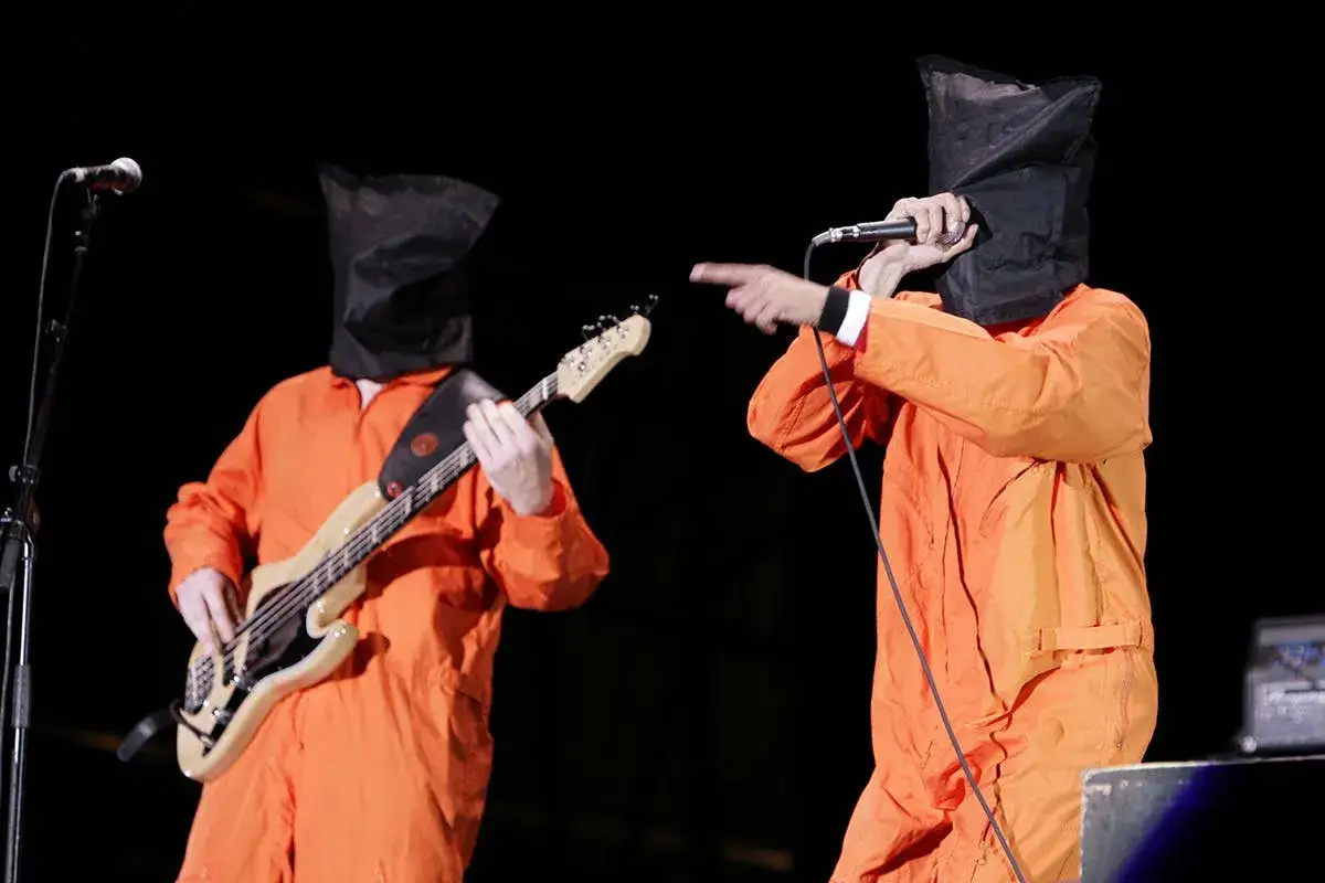 Zwei Musiker mit Maske, in orangefarbenen Overalls, performen auf der Bühne. Einer spielt Bass, der andere singt ins Mikrofon.