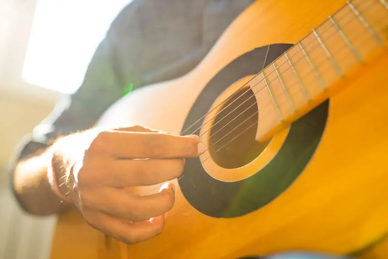 Eine Hand zupft sanft die Saiten einer Gitarre, w&auml;hrend Sonnenlicht durch das Schallloch f&auml;llt.