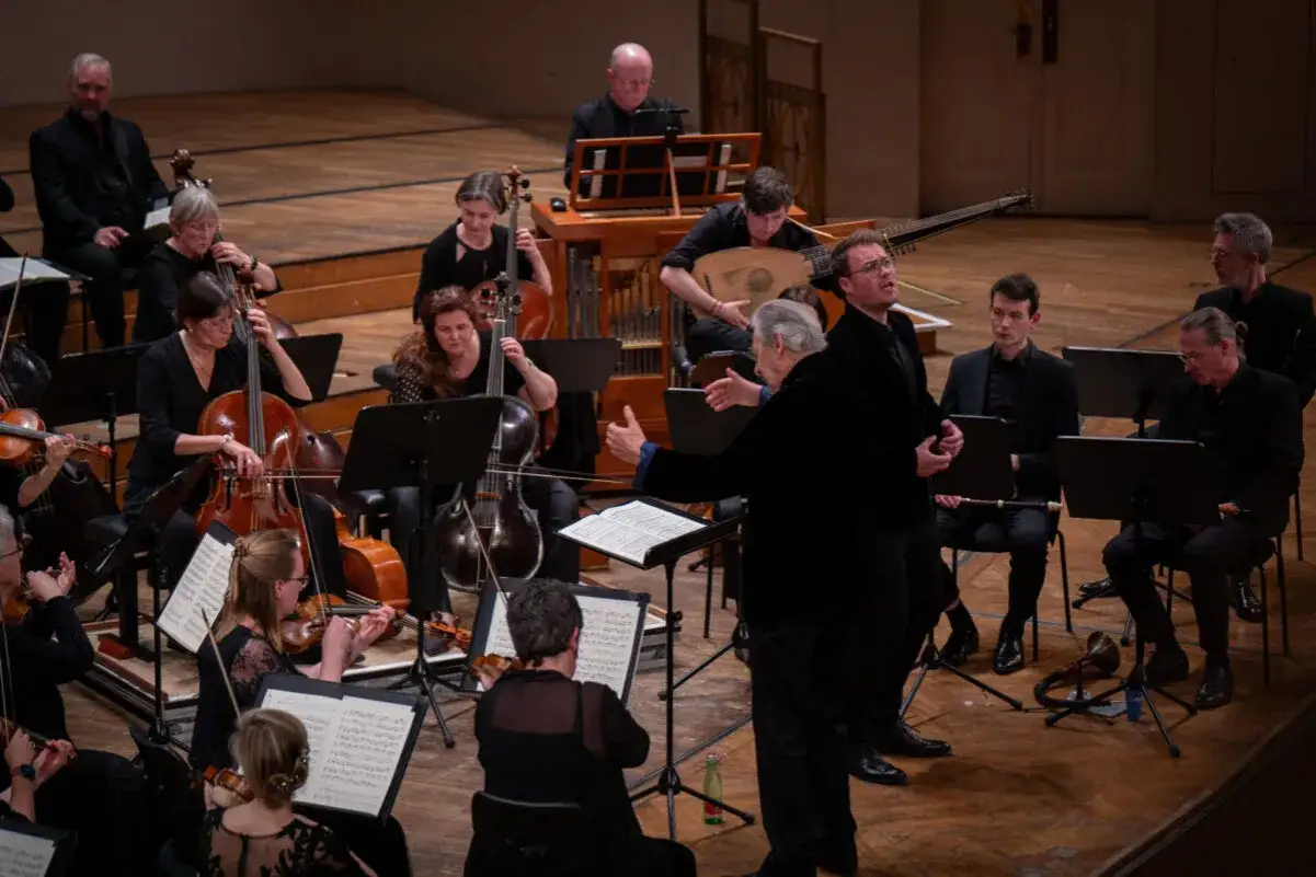 Ein Orchester spielt klassische Musik. Streicher und ein Dirigent sind zu sehen, im Hintergrund ein Cembalo und ein Lautenspieler.