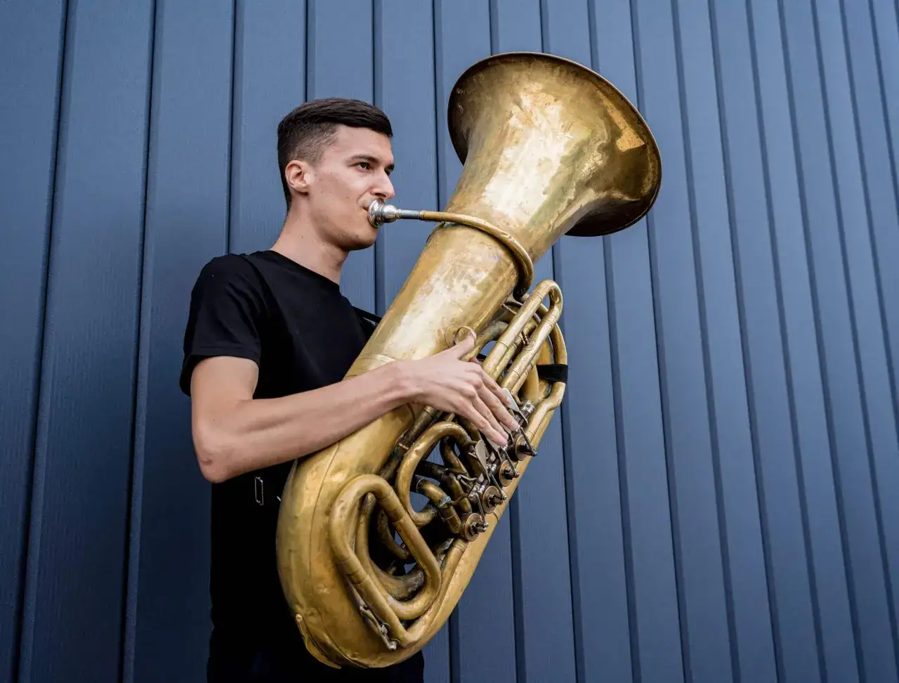Junger Mann spielt auf einer goldenen Tuba, einem beeindruckenden Blechblasinstrument, vor einer blauen Wand.