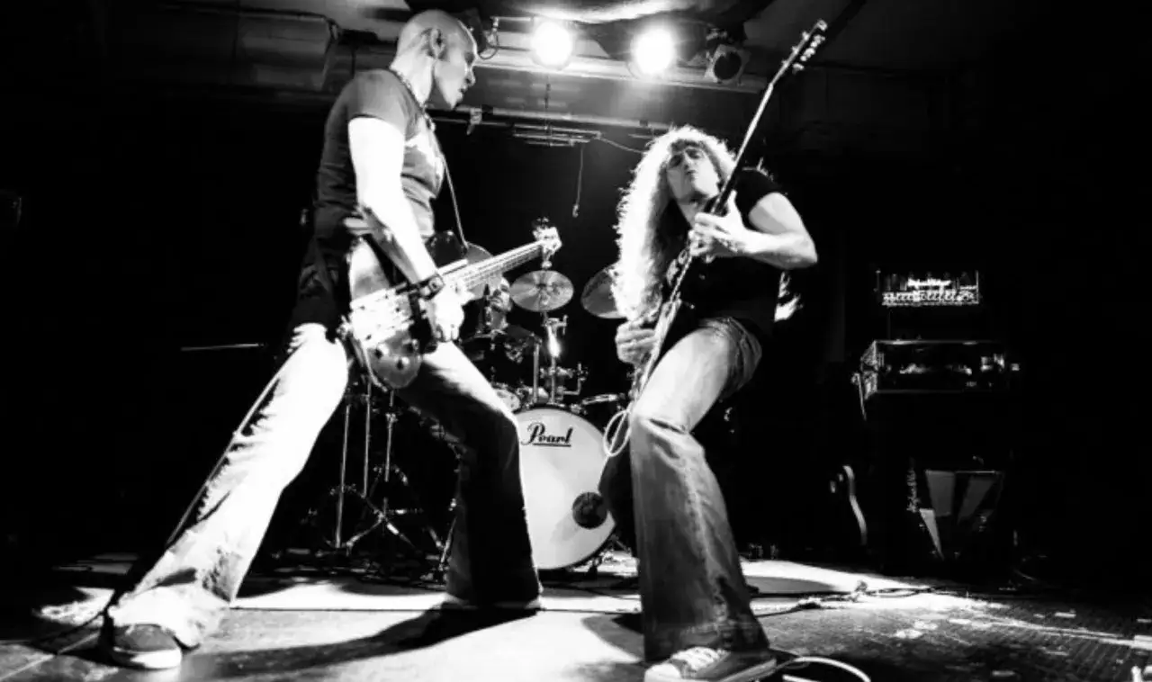 Zwei Rock-Musiker auf der Bühne, einer spielt Bass, der andere Gitarre. Schlagzeug im Hintergrund.