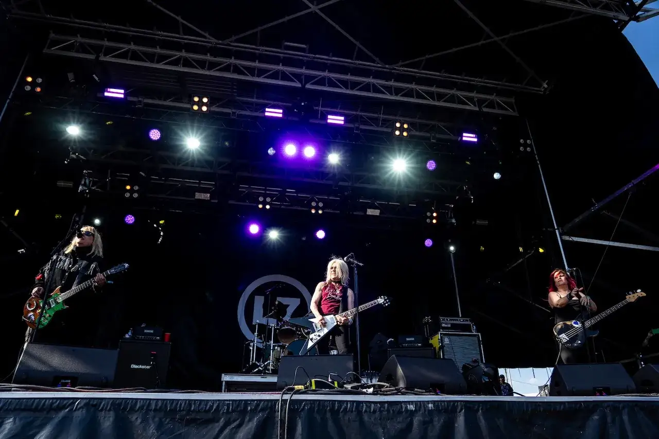 Drei Frauen einer Grunge-Band spielen auf einer Bühne unter bunten Lichtern.
