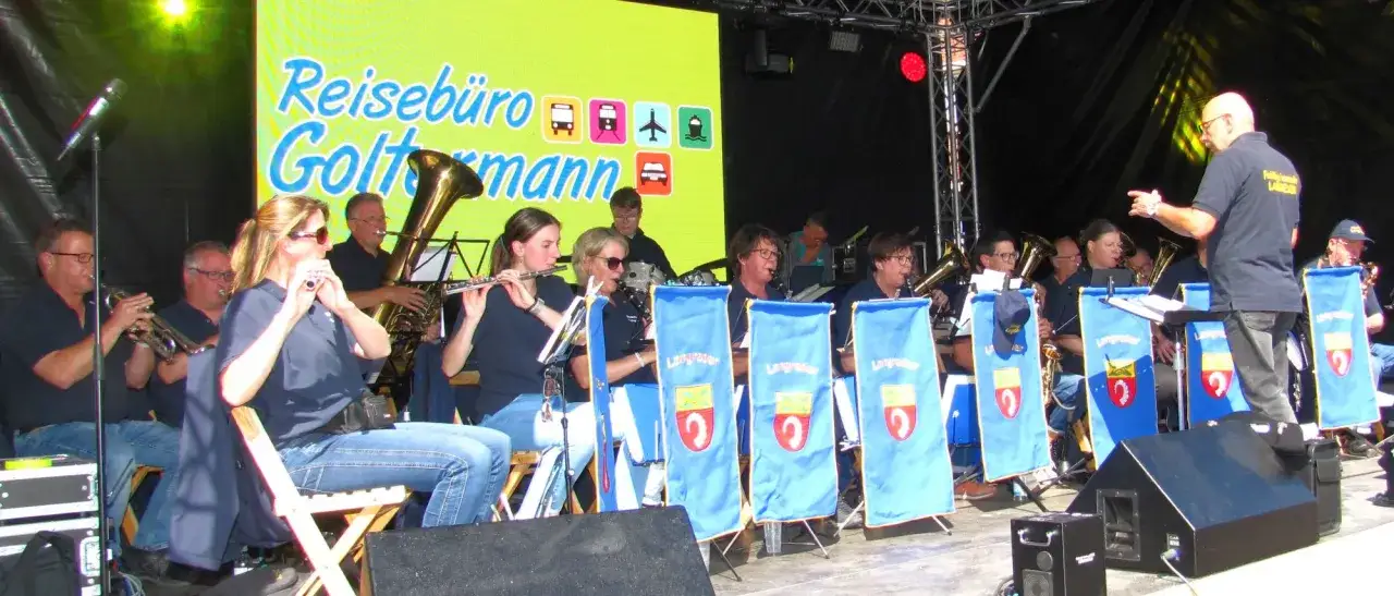 Eine Blaskapelle spielt auf einer Bühne vor einem bunten Banner. Die Musiker spielen verschiedene Instrumente, darunter Trompeten, Flöten und Tuben.
