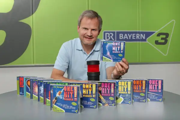 Mann präsentiert CDs mit dem Titel "One Hit Wonder". Viele weitere CDs dieser Reihe sind auf dem Tisch aufgereiht.