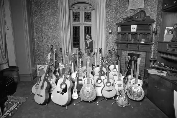 Mann mit vielen Rock n Roll Instrumenten, darunter Akustik- und E-Gitarren, vor einem Fenster.