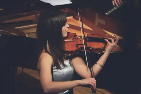 Eine Frau spielt Geige, ihr Bogen erzeugt einen zarten vibrato geige Klang. Im Hintergrund ist ein Klavier zu sehen.